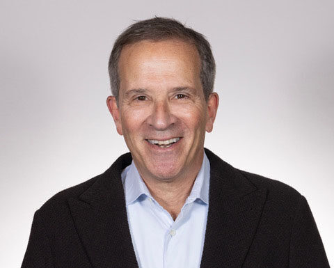 Smiling man in a light blue shirt and dark blazer against a neutral backdrop.