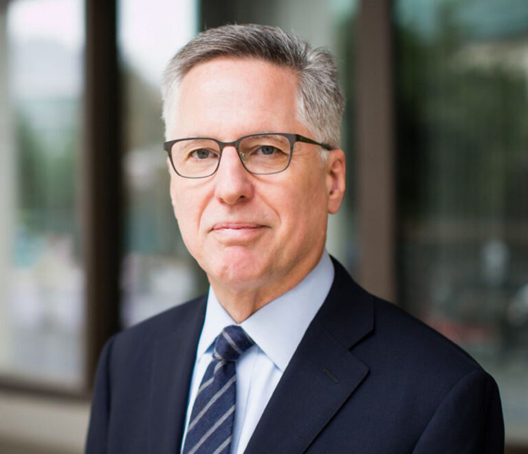 Head-and-shoulders portrait of a middle-aged man in a navy suit and striped tie, wearing glasses, outdoors near a glass building.