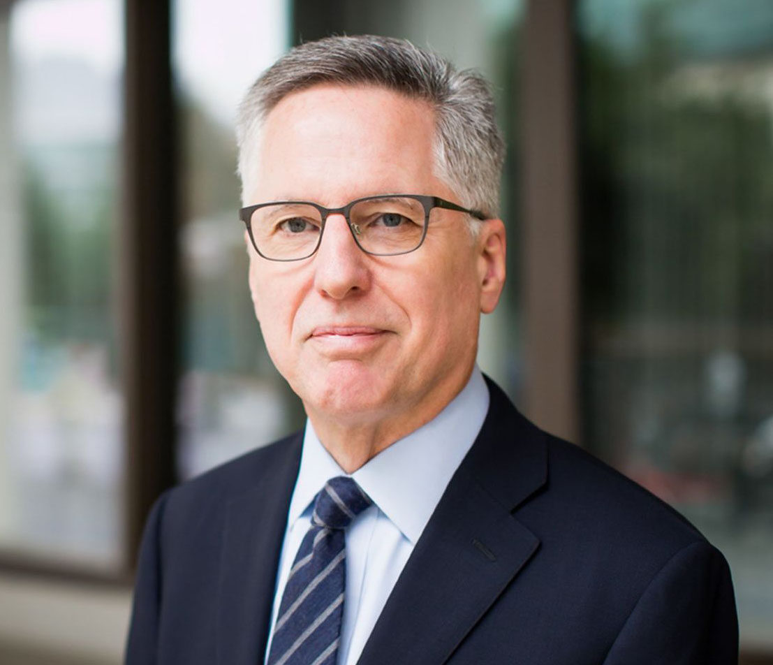 Head-and-shoulders portrait of a middle-aged man in a navy suit and striped tie, wearing glasses, outdoors near a glass building.